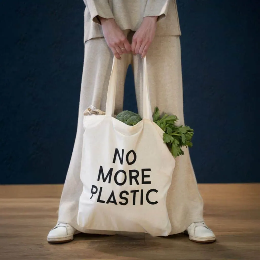 Sacola de tecido com mensagem "No More Plastic" contendo legumes e vegetais frescos, promovendo sustentabilidade e consumo consciente.